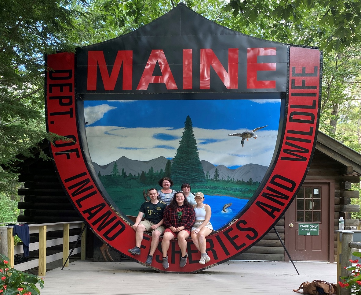 Maine Government Summer Internship students sitting on a large structure depicting the logo of the Maine Department of Inland Fisheries and Wildlife.