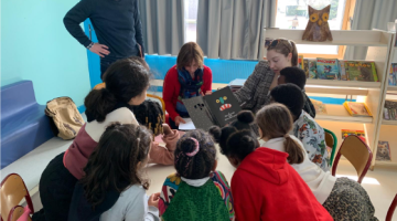 Group of children gathered around Nicole LeBlanc and a coworker reading a book in a classroom.