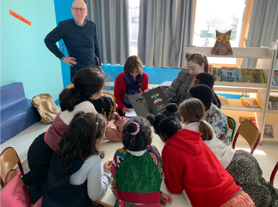 Group of children gathered around Nicole LeBlanc and a coworker reading a book in a classroom.