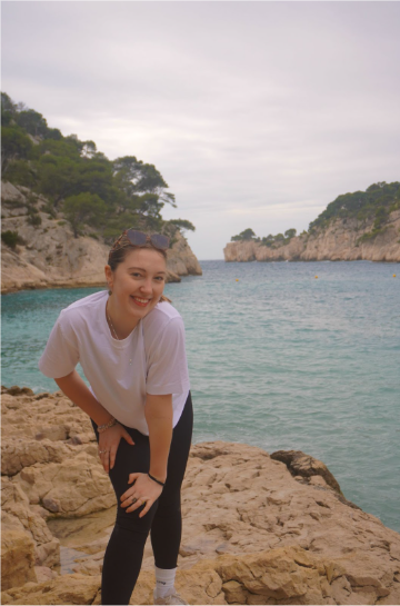 Nicole LeBlanc smiling on rocky shore with cliffs and sea in background.
