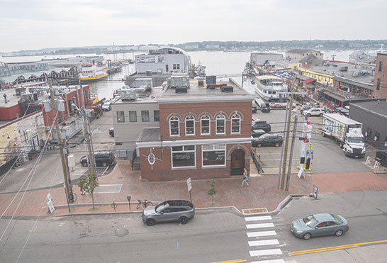 High-angle view of a red brick commercial building in a waterfront district with boats docked in the background harbor.