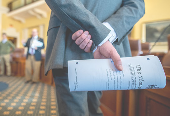 Close-up of a person in a suit holding a rolled legislative document behind their back inside a government chamber.
