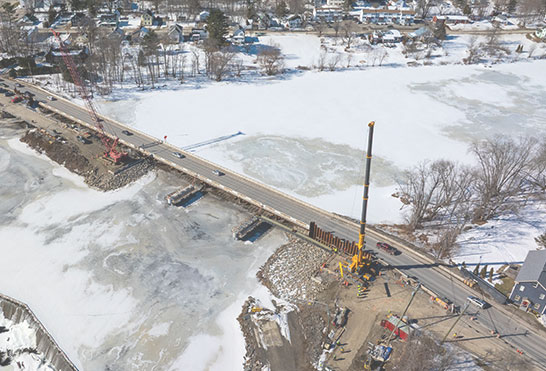 Aerial view of a bridge undergoing construction across a frozen, ice-covered body of water with tall cranes stationed on site.